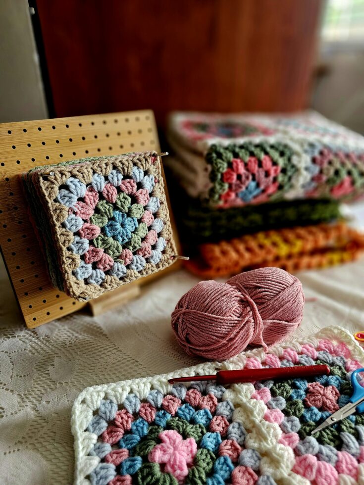 Pastel granny squares with yarn, a crochet hook, and scissors arranged on a soft cream bed