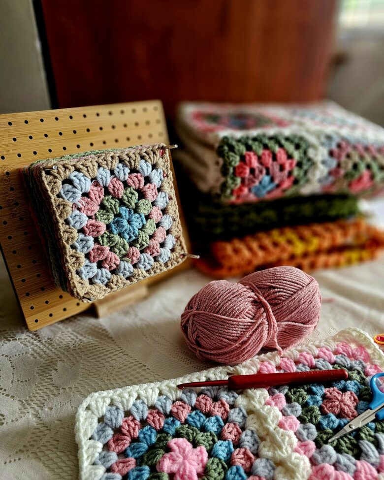 Pastel granny squares with yarn, a crochet hook, and scissors arranged on a soft cream bed